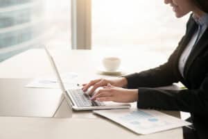 Mulher com roupa preta, teclando em um notebook cinza com folhas de apresentação sobre a mesa, com gráficos.